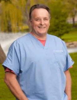Male chiropractor in blue scrubs smiling outdoors with blurred green background
