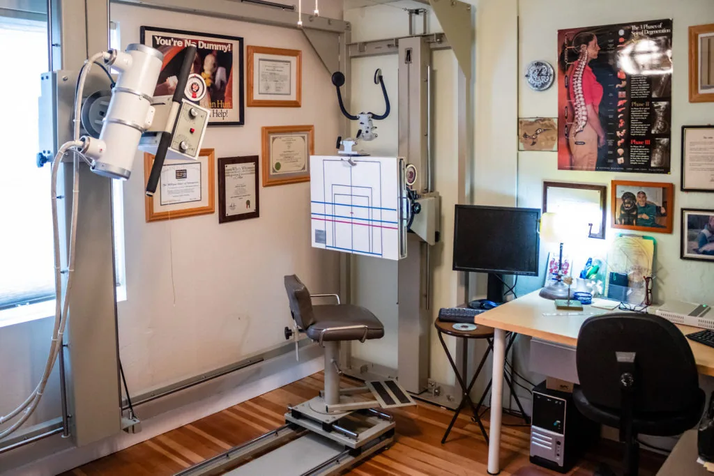 Chiropractic office with examination equipment, desk, computer monitor, and framed certifications and posters on walls