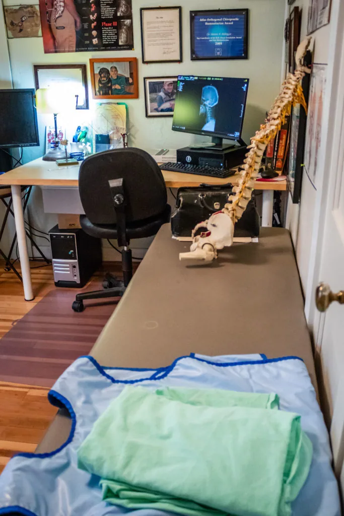 Home office and treatment room with desk, computer monitor, black office chair, skeleton model, framed certificates and photos on walls, and folded clothing in foreground