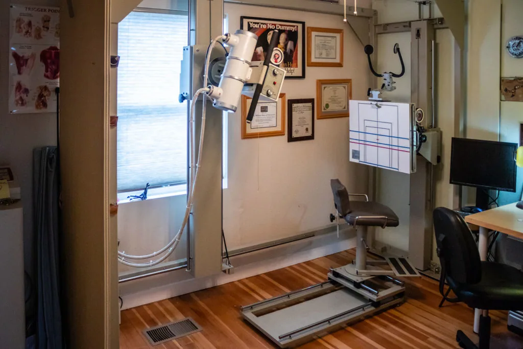 Chiropractic office treatment room with exercise equipment, framed certificates on wall, and water view through glass door