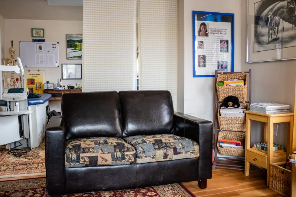 Black leather couch with patterned cushions in a chiropractic office waiting room with desk, storage furniture, and bulletin boards on walls