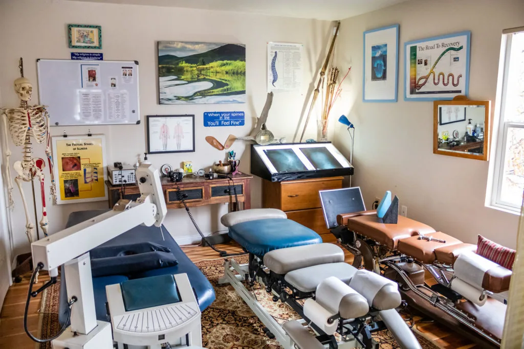 Chiropractic treatment room with adjustment tables, anatomical charts, computer workstations, and skeletal model