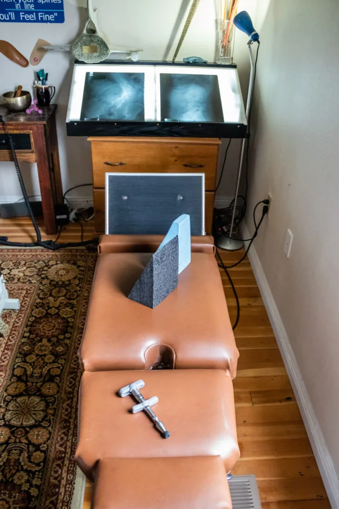 Chiropractic treatment room with orange massage table, overhead X-ray viewer displaying spinal images, and treatment equipment.