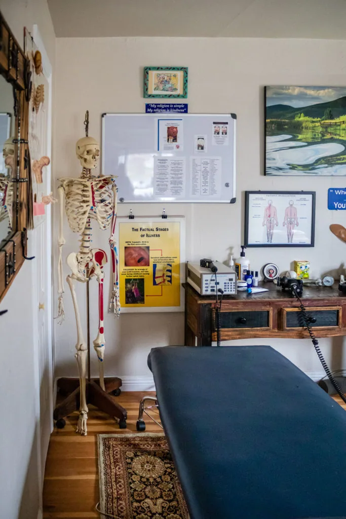 Chiropractic clinic room with anatomical skeleton model, treatment table, wall charts including muscular system diagram, whiteboard with notes, and medical equipment on desk