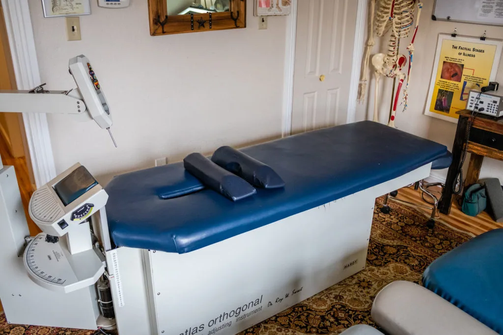 Chiropractic treatment table with blue padding and black bolsters in clinical examination room with anatomical charts and equipment
