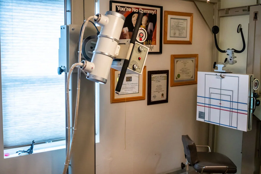 Chiropractic examination room with X-ray machine, framed certifications on wall, and treatment equipment