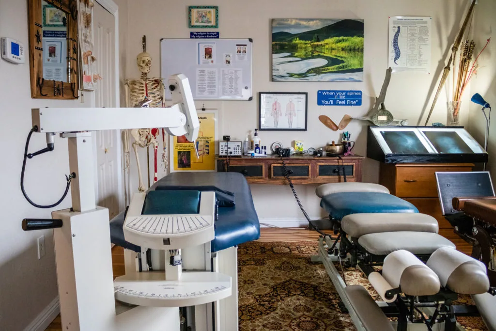 Chiropractic treatment room with examination table, adjustable chair, medical equipment, and anatomical charts on walls