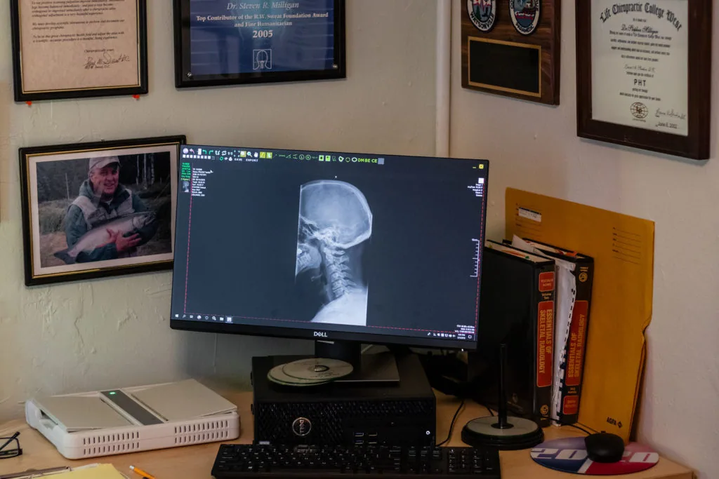 Office desk with medical imaging software displaying X-ray on monitor, surrounded by framed certificates and photos on walls