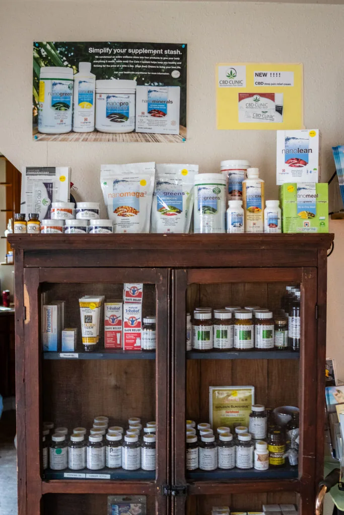 Wooden display cabinet with glass doors containing nutritional supplement bottles and containers, with additional products displayed on top shelf and promotional posters on wall above