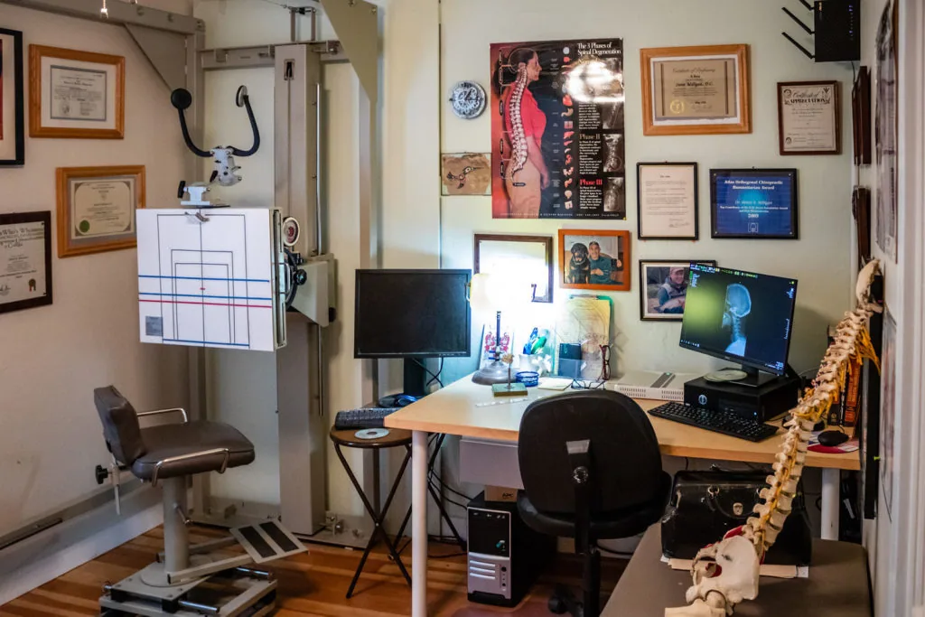 Chiropractic office workspace with desk, computer monitors, medical equipment, framed certificates on walls, and anatomical models
