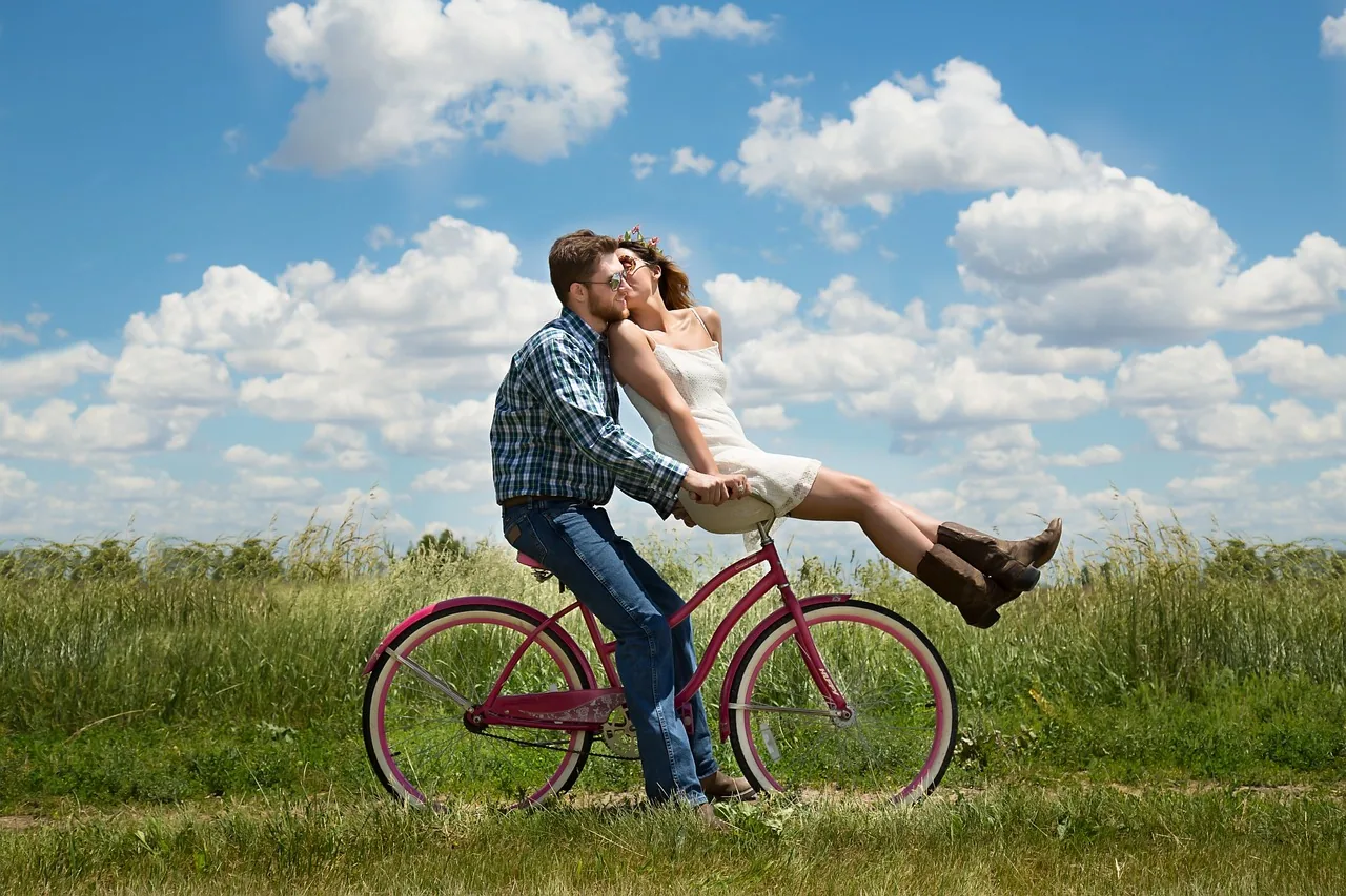 Man in checkered shirt and woman in white top riding together on purple bicycle through grassy field under blue sky with white clouds