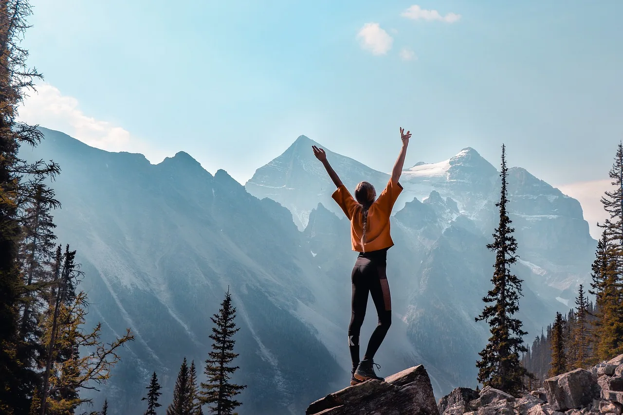 Person in yellow jacket standing on rock with arms raised celebrating, overlooking mountain range with evergreen trees